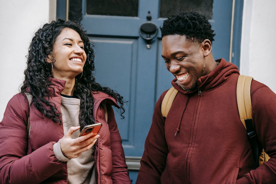 Multiethnic happy students laughing while using mobile phone near door of building in daytime