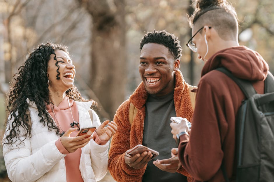Cheerful multiracial friends in trendy outfits laughing while browsing mobile phones and communicating with each other