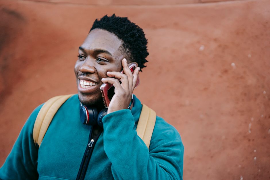Cheerful young African American male student in casual clothes and backpack smiling and looking away while having phone conversation near old stone building